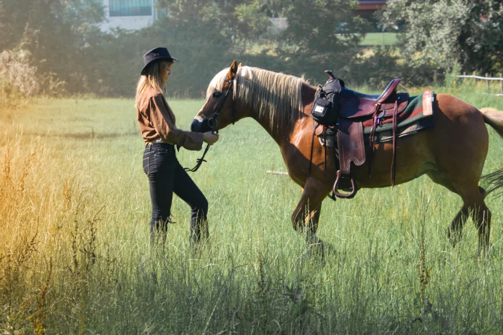 A woman wearing a cowboy hat and brown shirt stands in a grassy field, gently holding the reins of a saddled palomino horse.